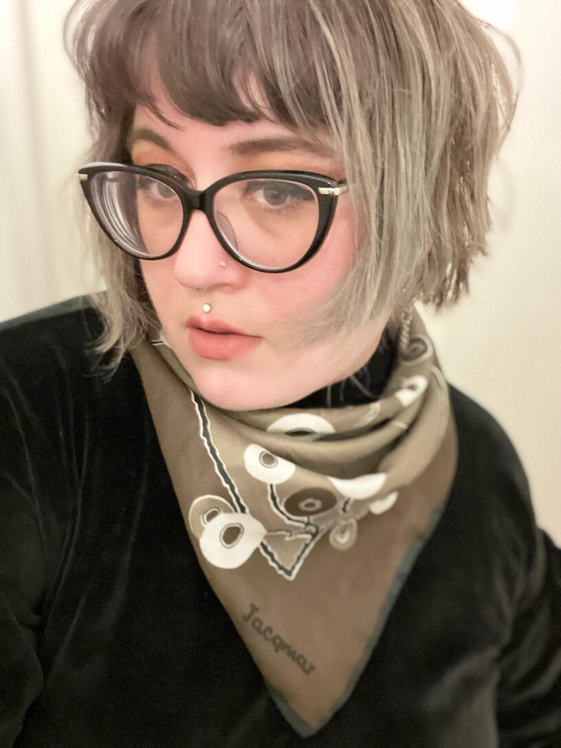 Portrait photo of a person with a grey bob wearing large glasses, a black top and a vintage scarf